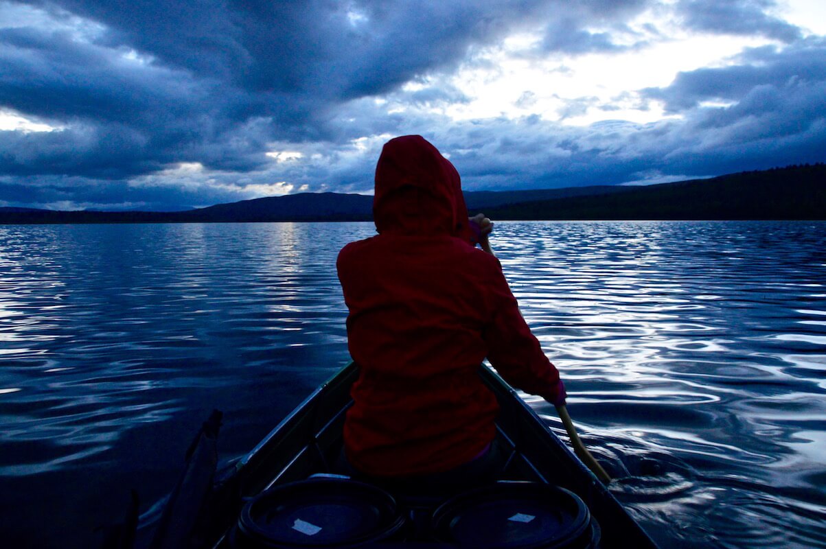 Sophie in her red Fjällräven jacket paddling out into the deep blue lake in front of the canoe at midnight in the Swedish summer