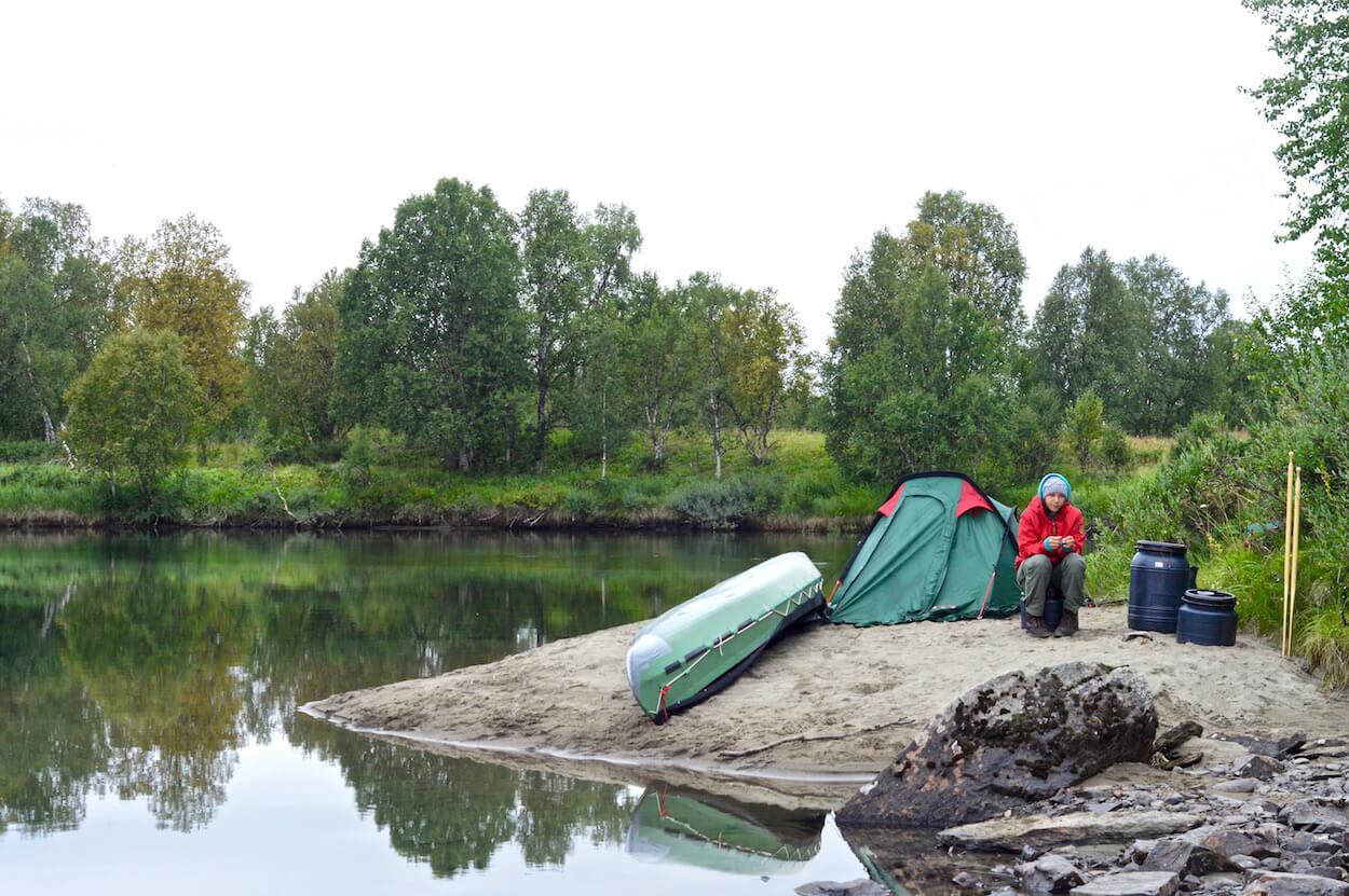 Sophie sitting on a small sandy island right next to our Ally canoe and our tiny tent in Lapland, Sweden