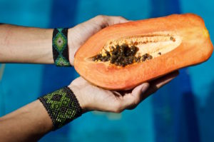 Papaya held by two hands over a swimming pool with two Mexican bracelets on the wrist